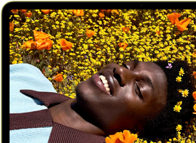 MacBook Neo, color amarillo cítrico, la pantalla muestra a una persona sonriente acostada en un campo de flores bajo el sol